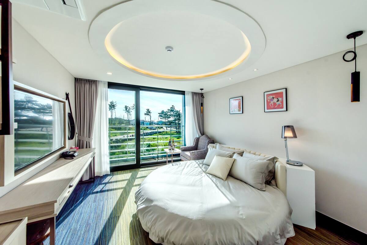 A modern hotel room with a large bed, a desk, a chair, a nightstand, and a view of a green landscape through the window.