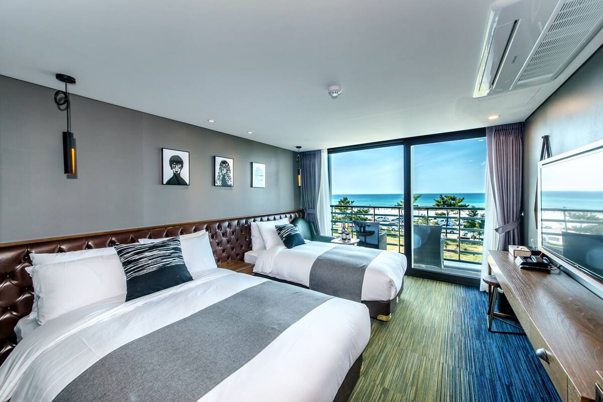 A hotel room with two beds, a large window with a view of the sea, and a desk with a chair.