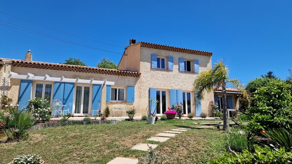 A two-story house with a tiled roof, blue shutters, and a garden with various plants and flowers.