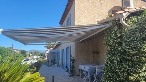 Un espacio al aire libre cubierto con una mesa, sillas, una planta en maceta y un edificio con contraventanas azules.