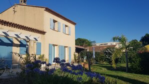 Una casa de dos pisos con contraventanas azules y tejado de tejas, rodeada de un jardín cuidado con diversas plantas y flores.