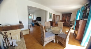 A spacious living area with a glass-top dining table surrounded by wicker chairs, a television, and a large wooden cabinet.