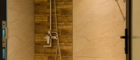 A shower with a handheld showerhead and a wall-mounted faucet, set against a brick wall and a marble-tiled wall.