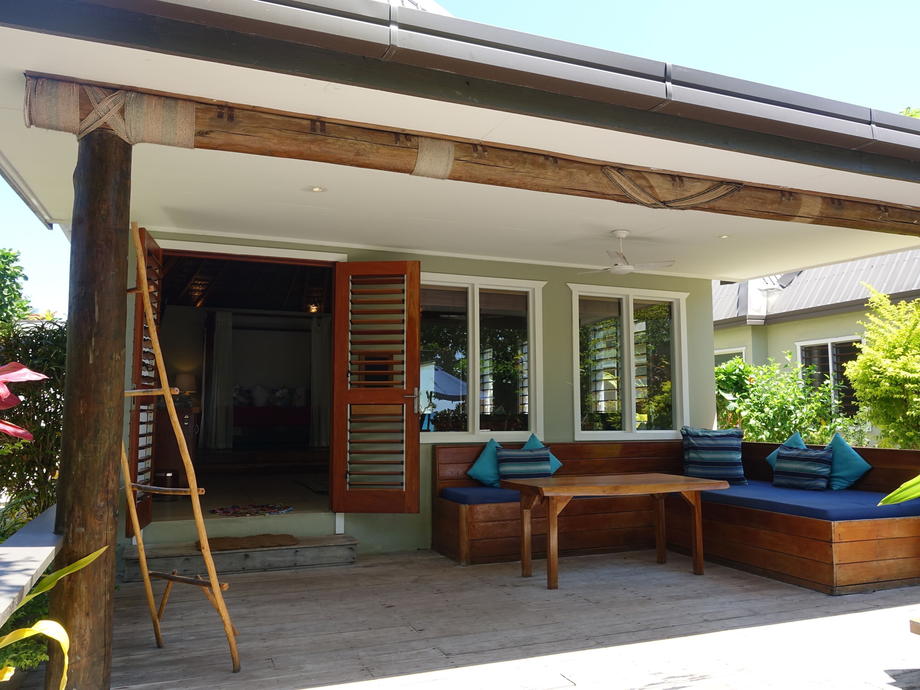 A covered outdoor seating area with a wooden bench and table, a ladder, and a wooden door leading to an interior space.