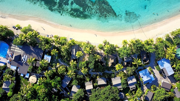 Luchtbeeld van een strandresort met helder turquoise water, een zandstrand en rijen huisjes in bungalowstijl, omgeven door palmbomen.