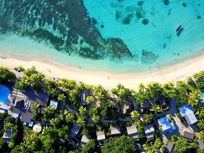 An aerial view of a beach resort with clear turquoise waters, a sandy beach, and rows of bungalow-style houses surrounded by palm trees. - Blue Lagoon Beach Resort (Nacula Island)