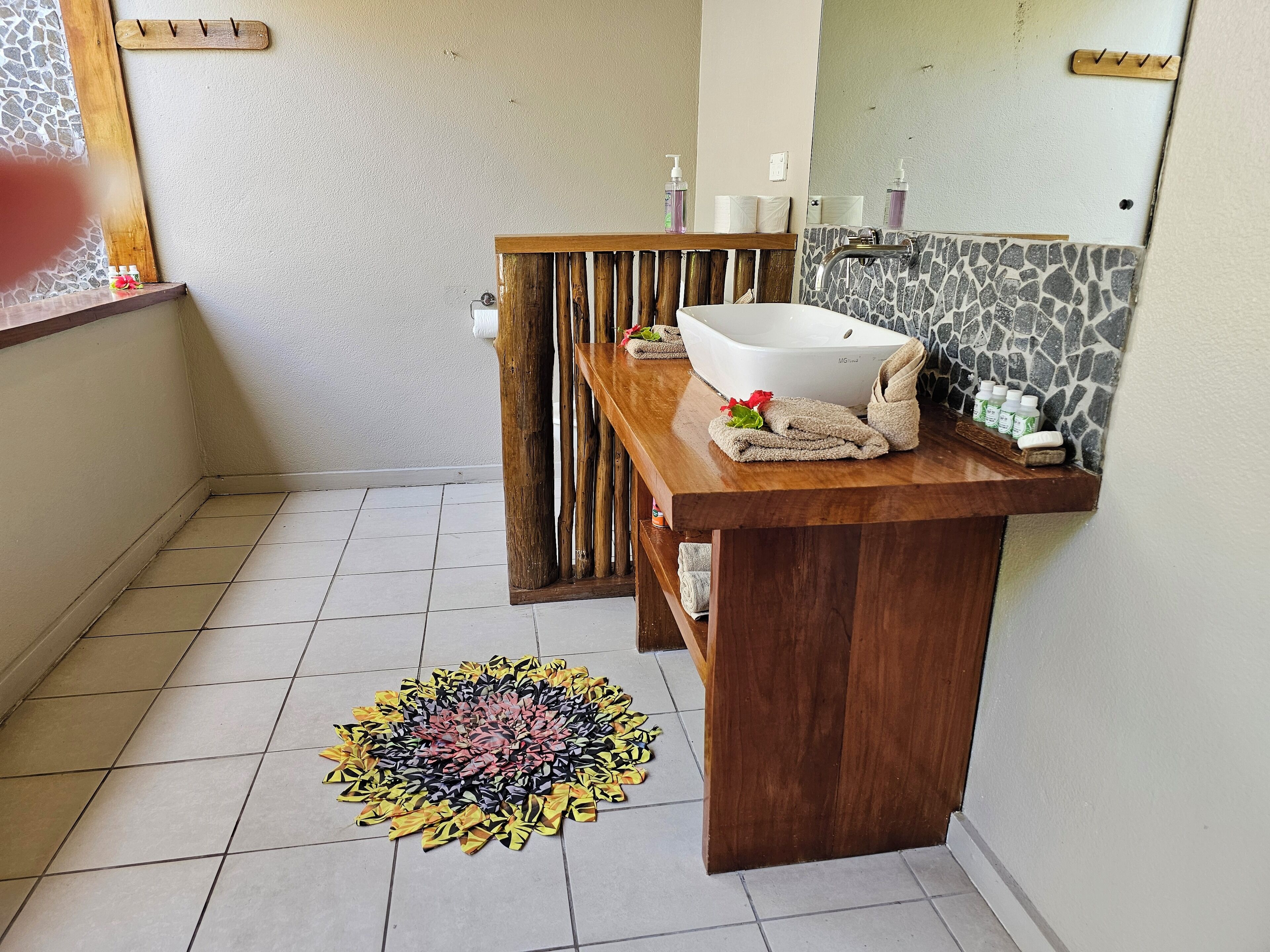 A bathroom with a wooden vanity, a white sink, and a mirror. There is a tiled floor and a decorative rug.