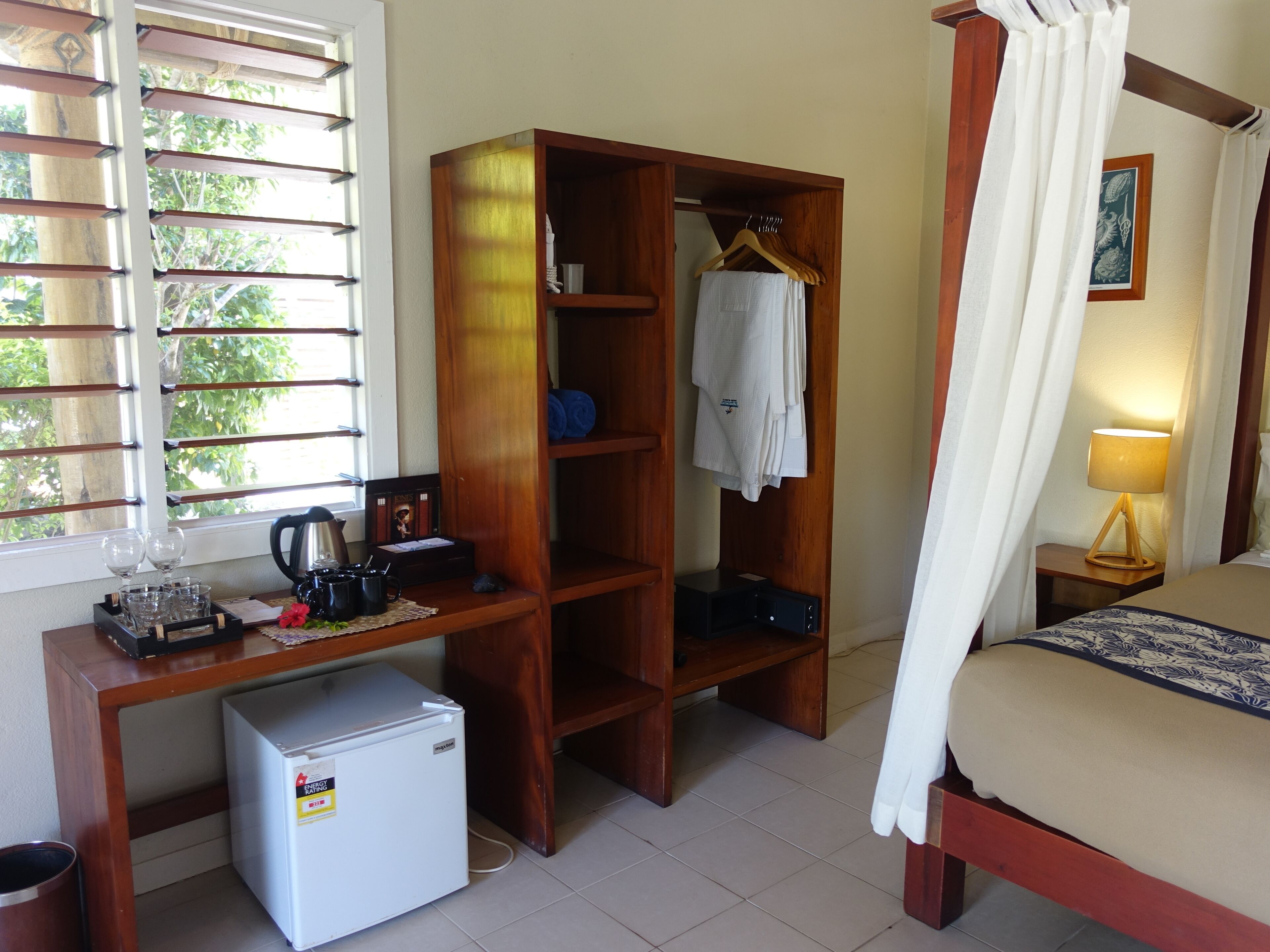 A bedroom with a bed, a wardrobe, a desk with a refrigerator, a window with blinds, and a bedside table with a lamp.