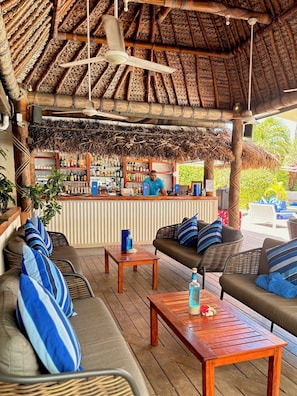 Un área de bar al aire libre con techo de paja, mobiliario de madera y una barra repleta de bebidas.