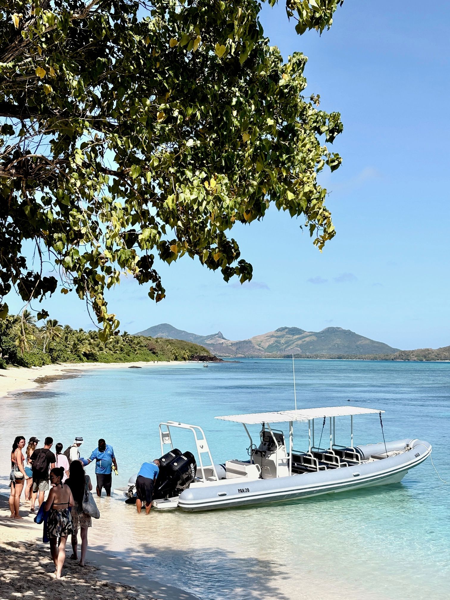 A beach with clear water, a boat, and people walking.