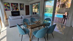 Een moderne woonkamer met een glazen eettafel, blauwe stoelen, een witte bank en grote ramen met uitzicht op een zwembad.