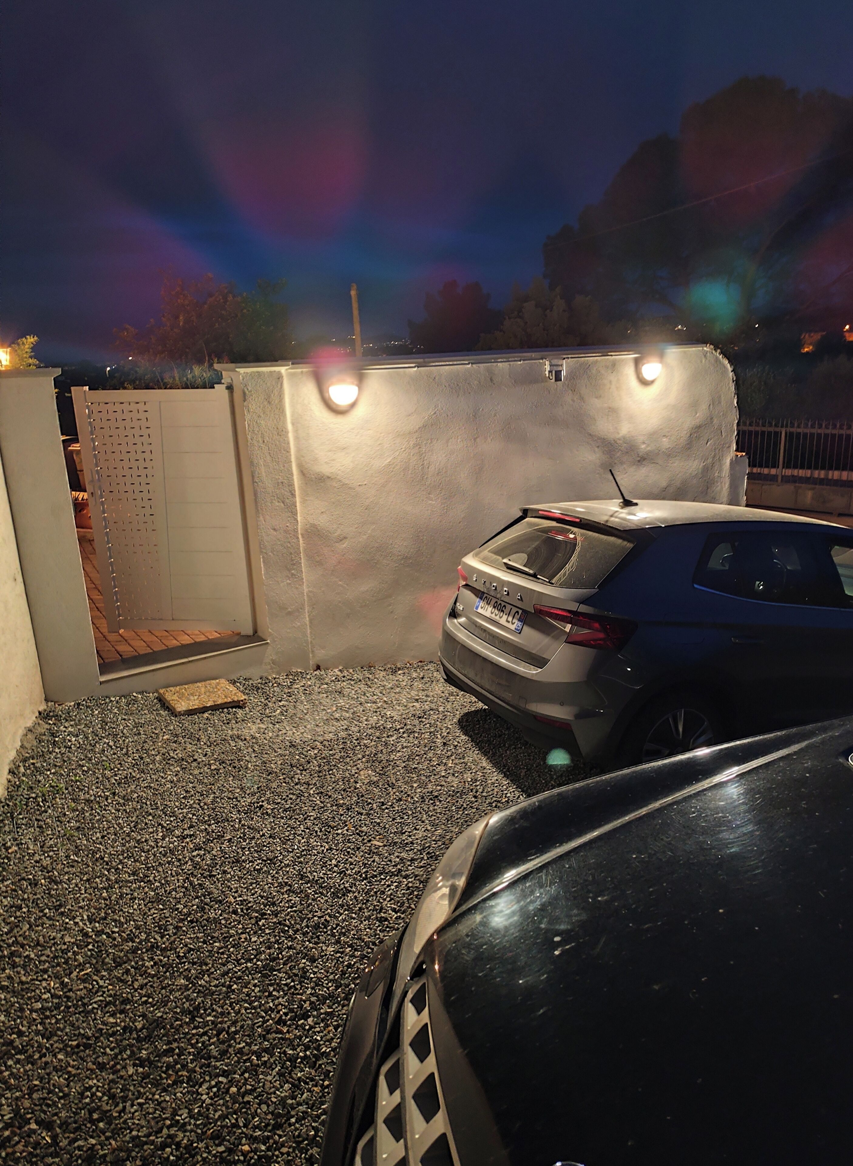 A parking area with a gravel surface, a car, and a wall with a gate.