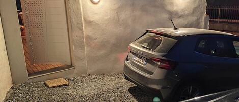 A parking area with a gravel surface, a car, and a wall with a gate.