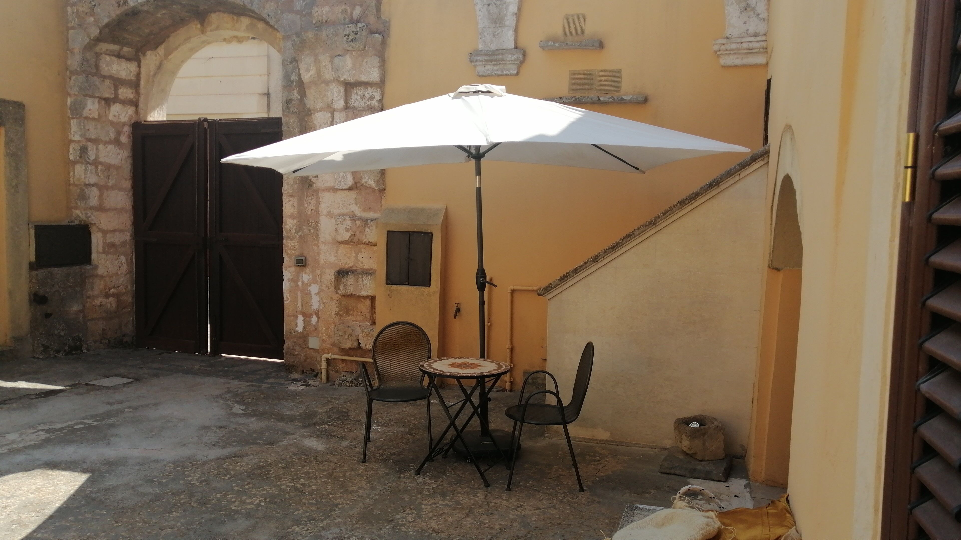An outdoor seating area with a table and chairs under a white umbrella, a stone wall, and a closed wooden gate.