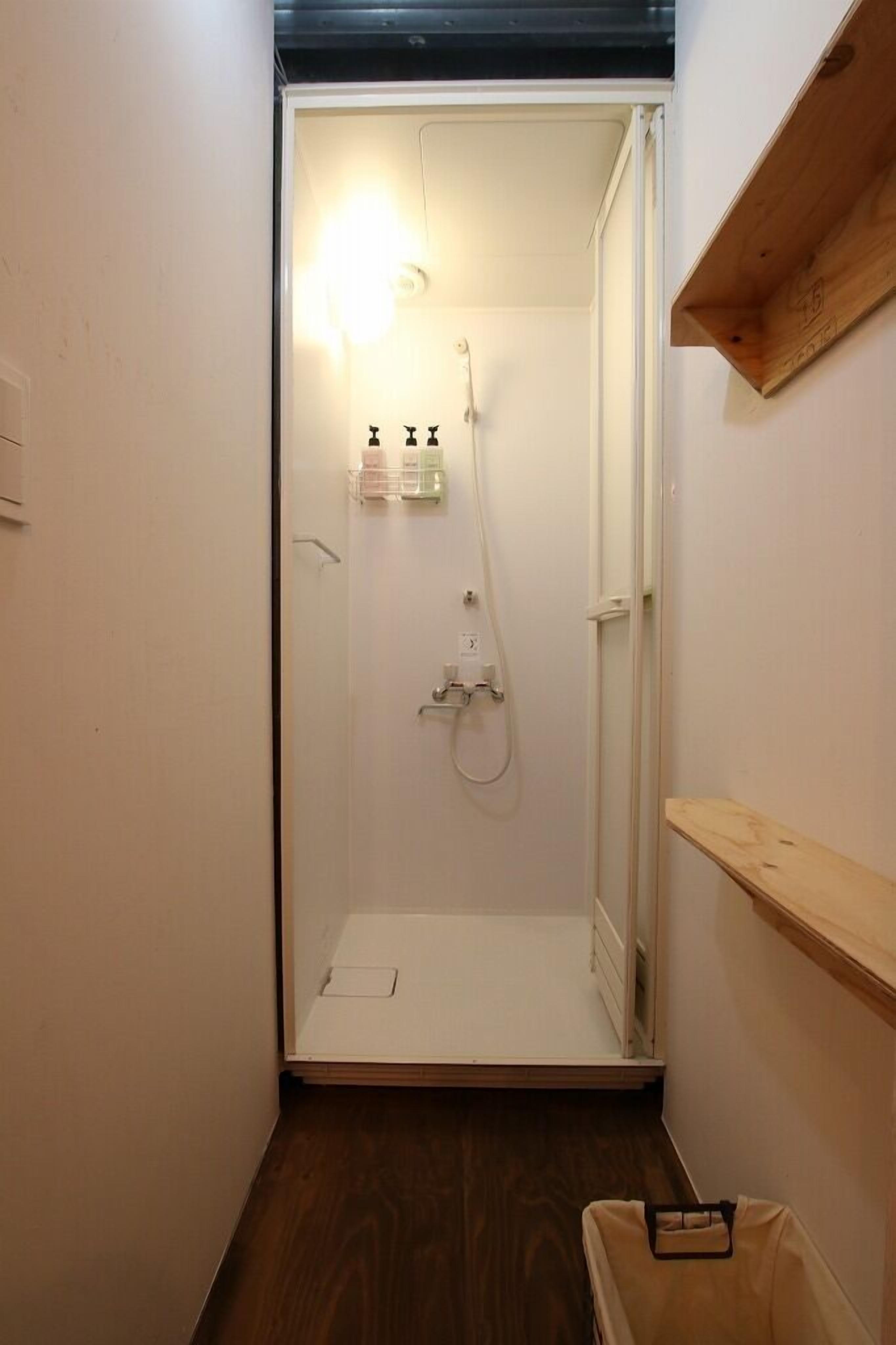 Un piccolo box doccia chiuso da una porta bianca, con uno scaffale in legno sopra e pavimento in legno.
