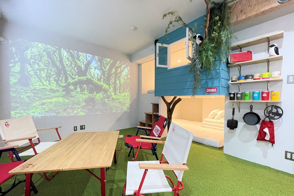 A room with a projector displaying a forest scene, a bunk bed with a hanging plant, a shelf with colorful containers, and a wall-mounted kitchen utensil rack.