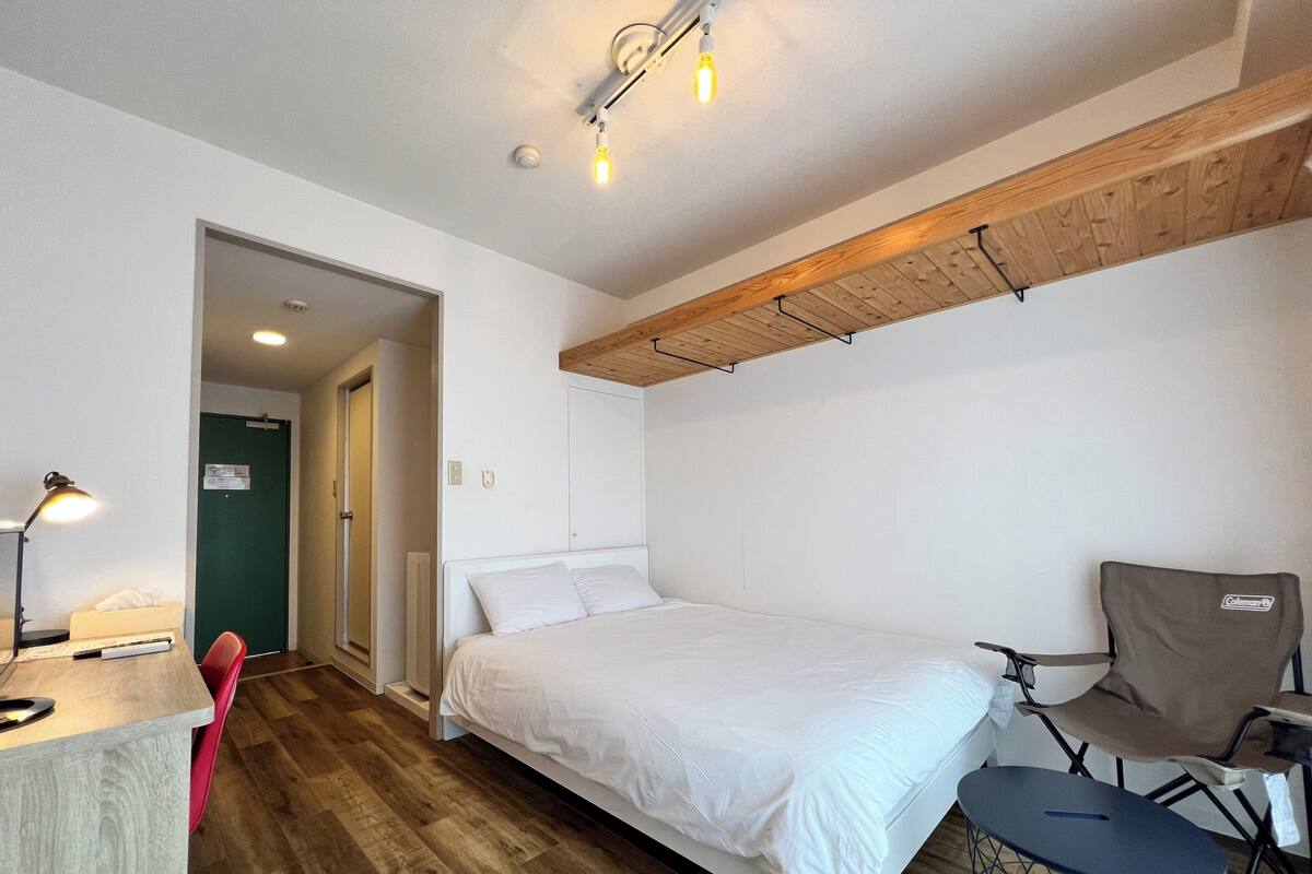 A modern hotel room with a wooden floor, a bed with white linens, a desk with a lamp, and a chair.