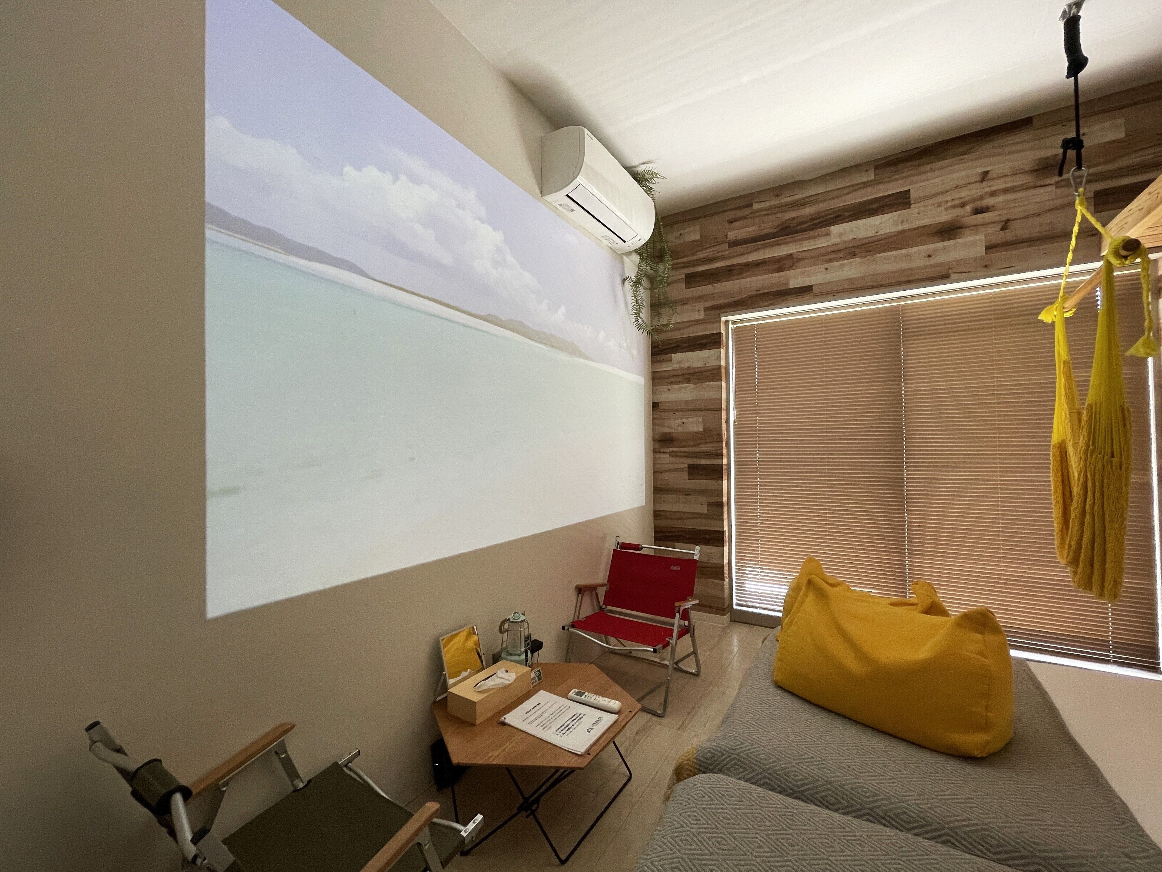 Una habitación con una pantalla de proyector mostrando una escena de playa, una pared de madera, un sillón de peluche amarillo y una silla roja.