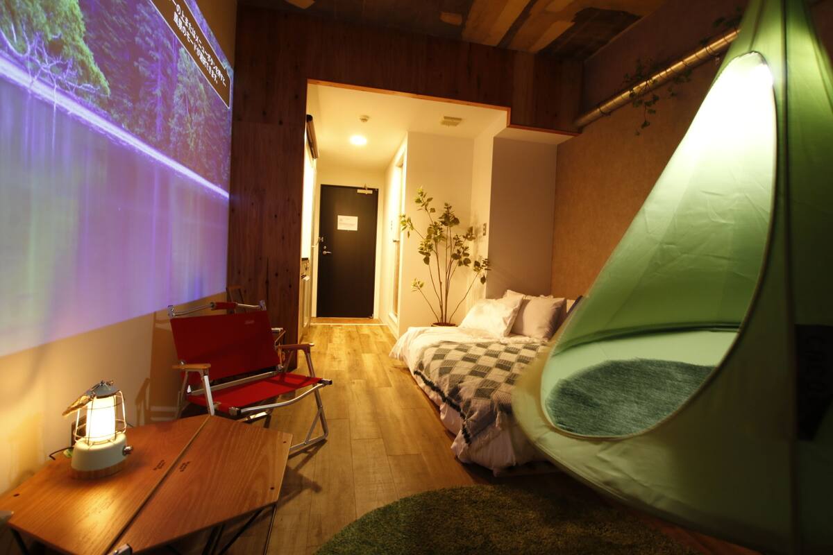 A room with a bed, a green tent-like structure, a projector screen, a red chair, and a table with a lantern.