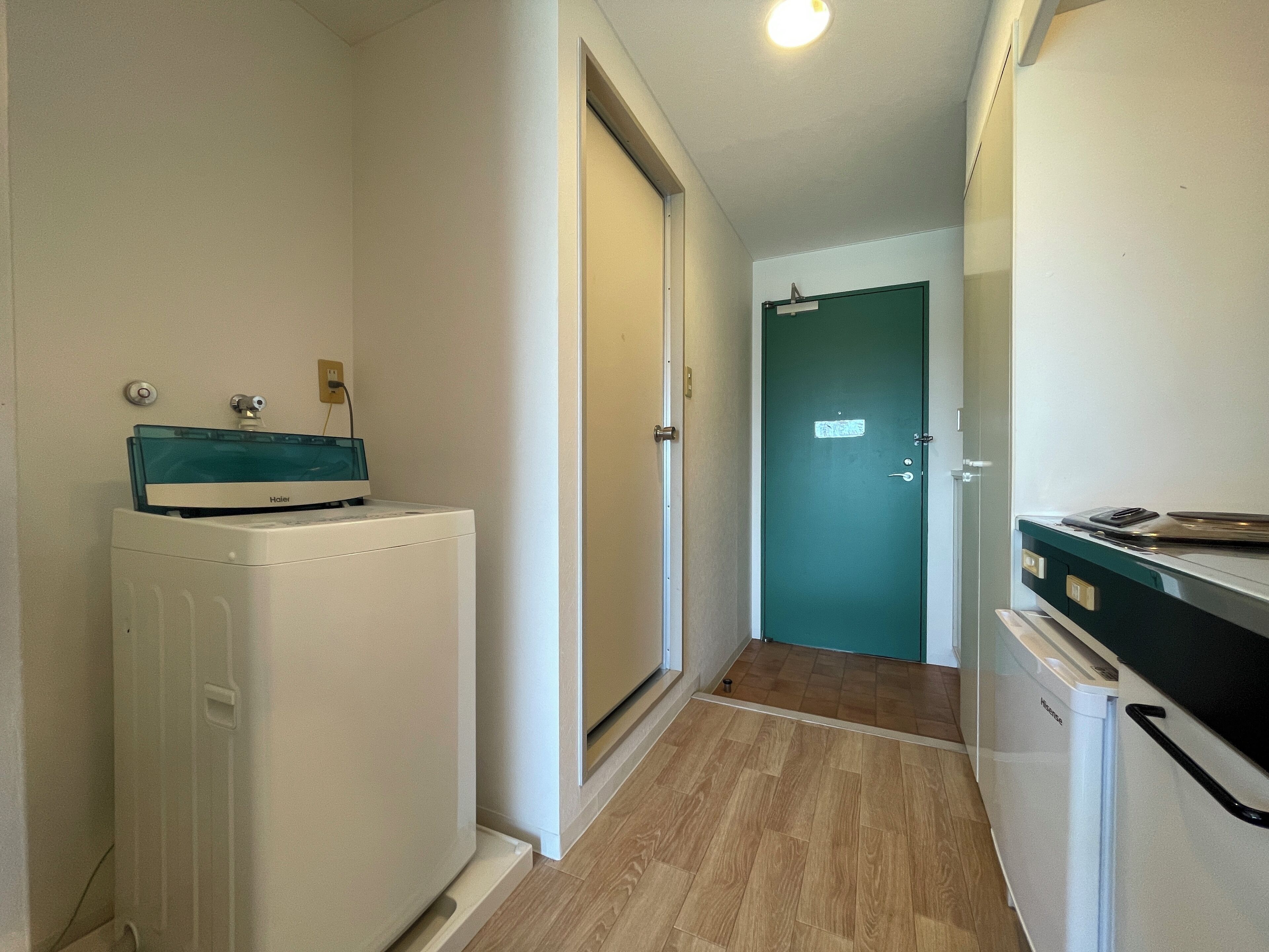 A narrow hallway with a washing machine, a small refrigerator, and a green door at the end.
