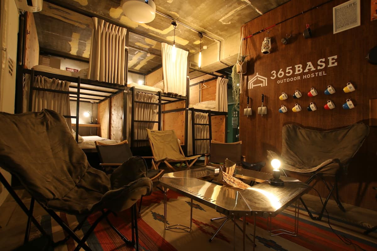A cozy hostel room with bunk beds, a table with a lamp, and a wall displaying various locks.
