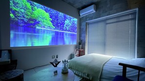 Un dormitorio con cama, una mesita de noche, una pantalla de proyector mostrando una escena natural, una ventana con persianas y un equipo de aire acondicionado.