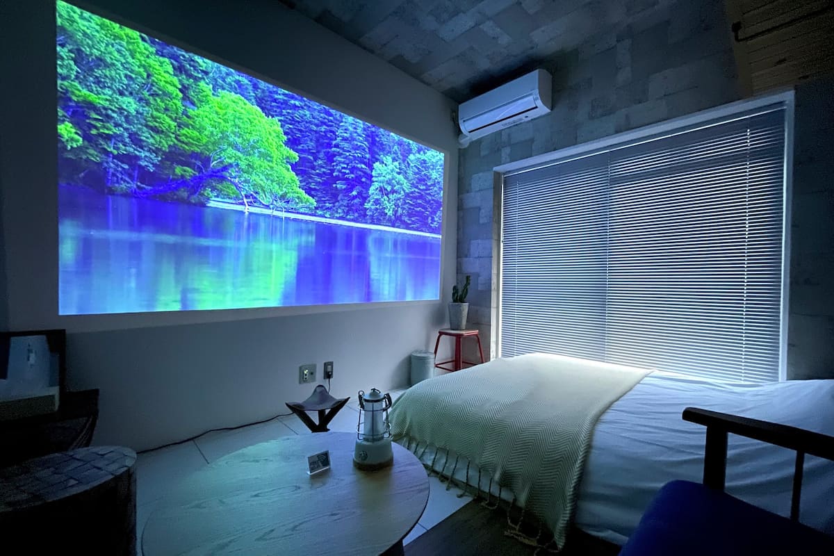 A bedroom with a bed, a bedside table, a projector screen displaying a nature scene, a window with blinds, and an air conditioning unit.