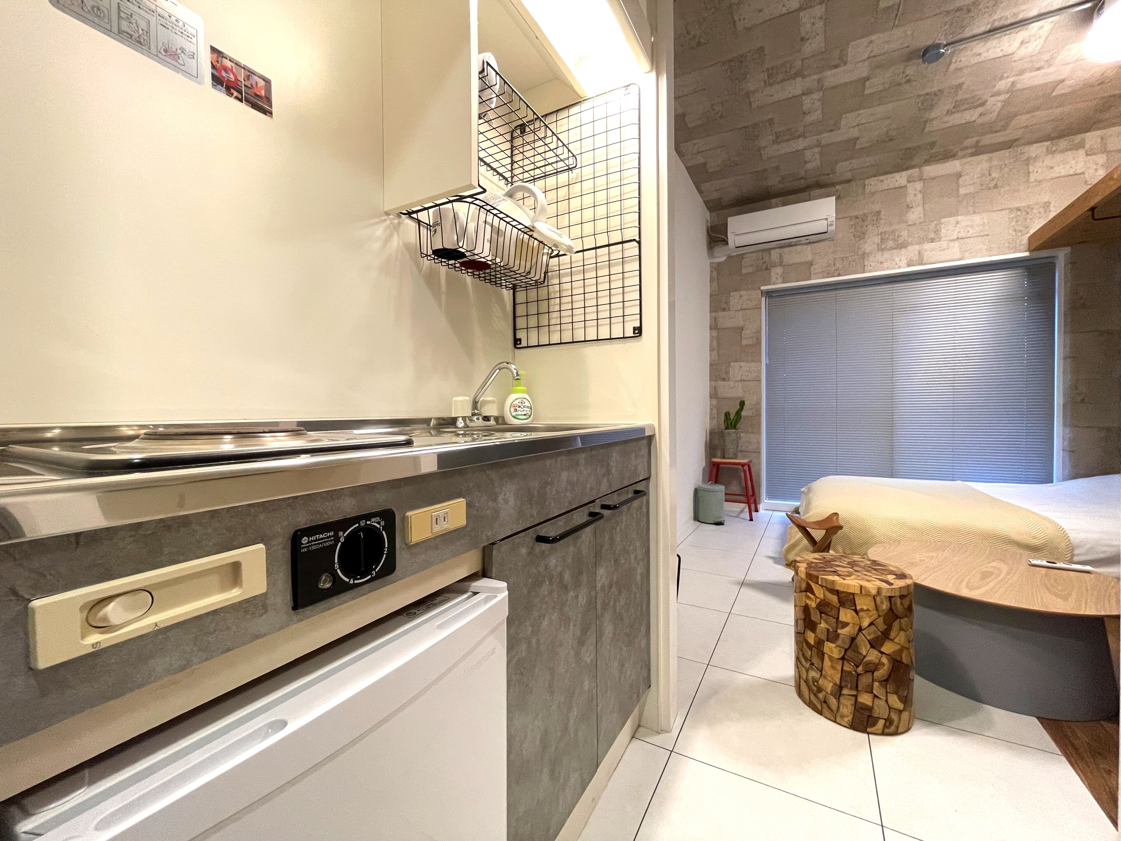 A compact kitchen with a built-in oven, a sink, and a microwave. There is a small wooden stool and a window with blinds in the adjacent room.