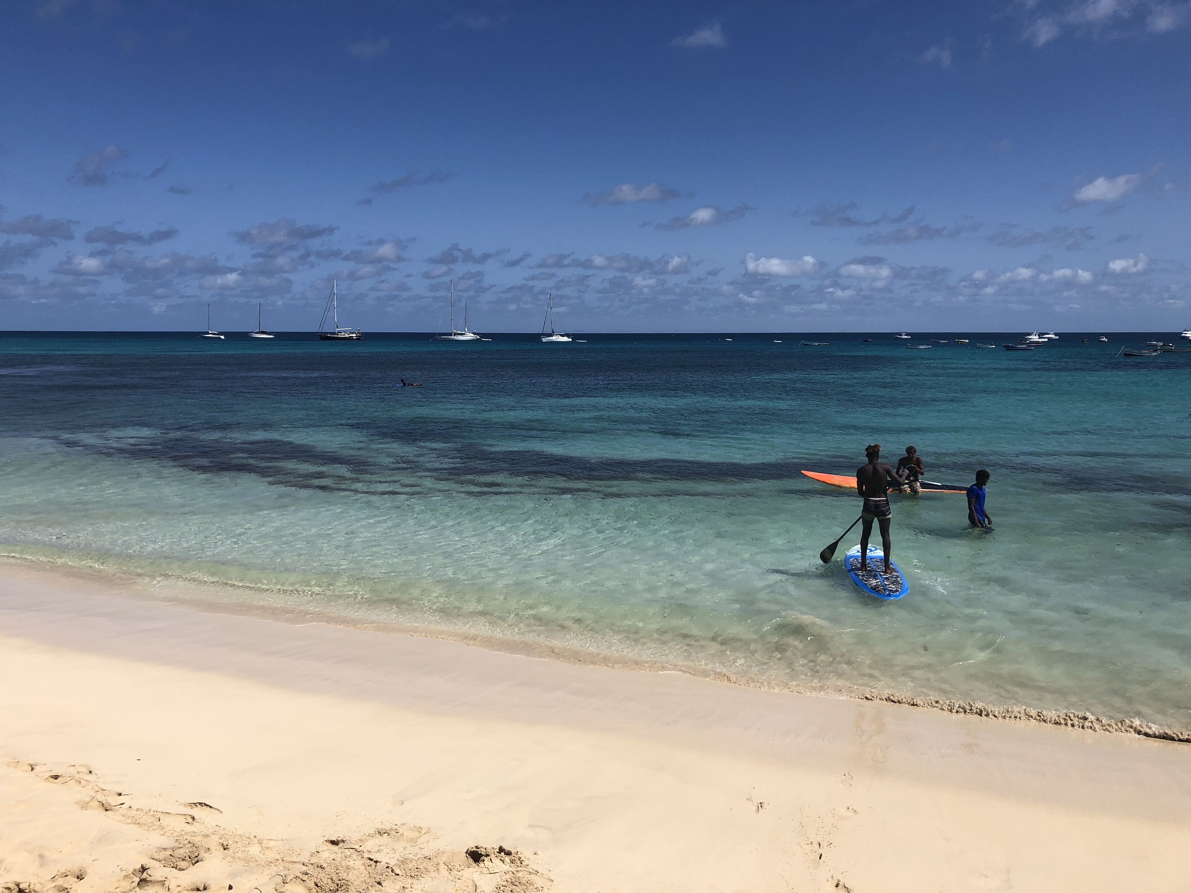 A beach with clear turquoise water, a sandy shore, and several boats in the distance.