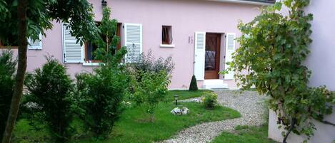 A pink house with white shutters, a gravel path, and green shrubs.