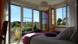 A bedroom with a large bed, a chair, and a view of greenery through open windows.