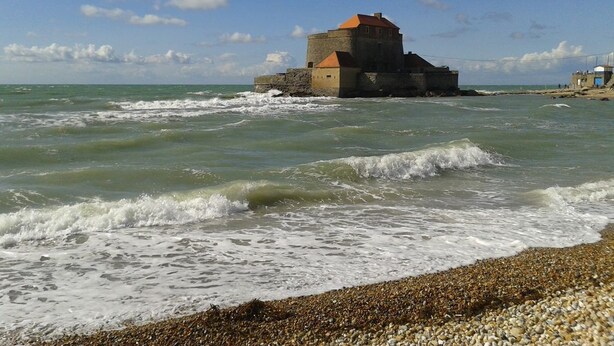 Une fortification côtière dotée d’une tour ronde, entourée de vagues et de galets.