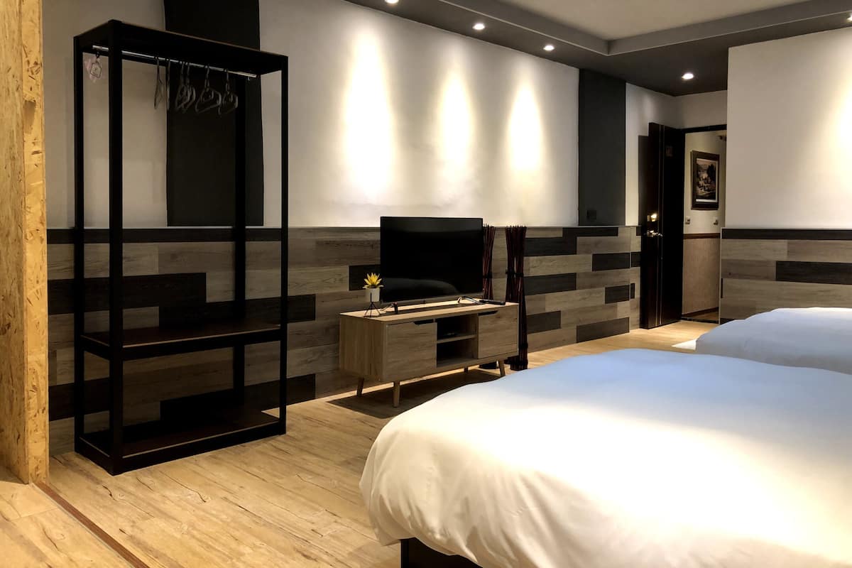 A modern hotel room with a wooden floor, a TV on a wooden stand, a bed with white bedding, and a wall-mounted shelf with hanging items.