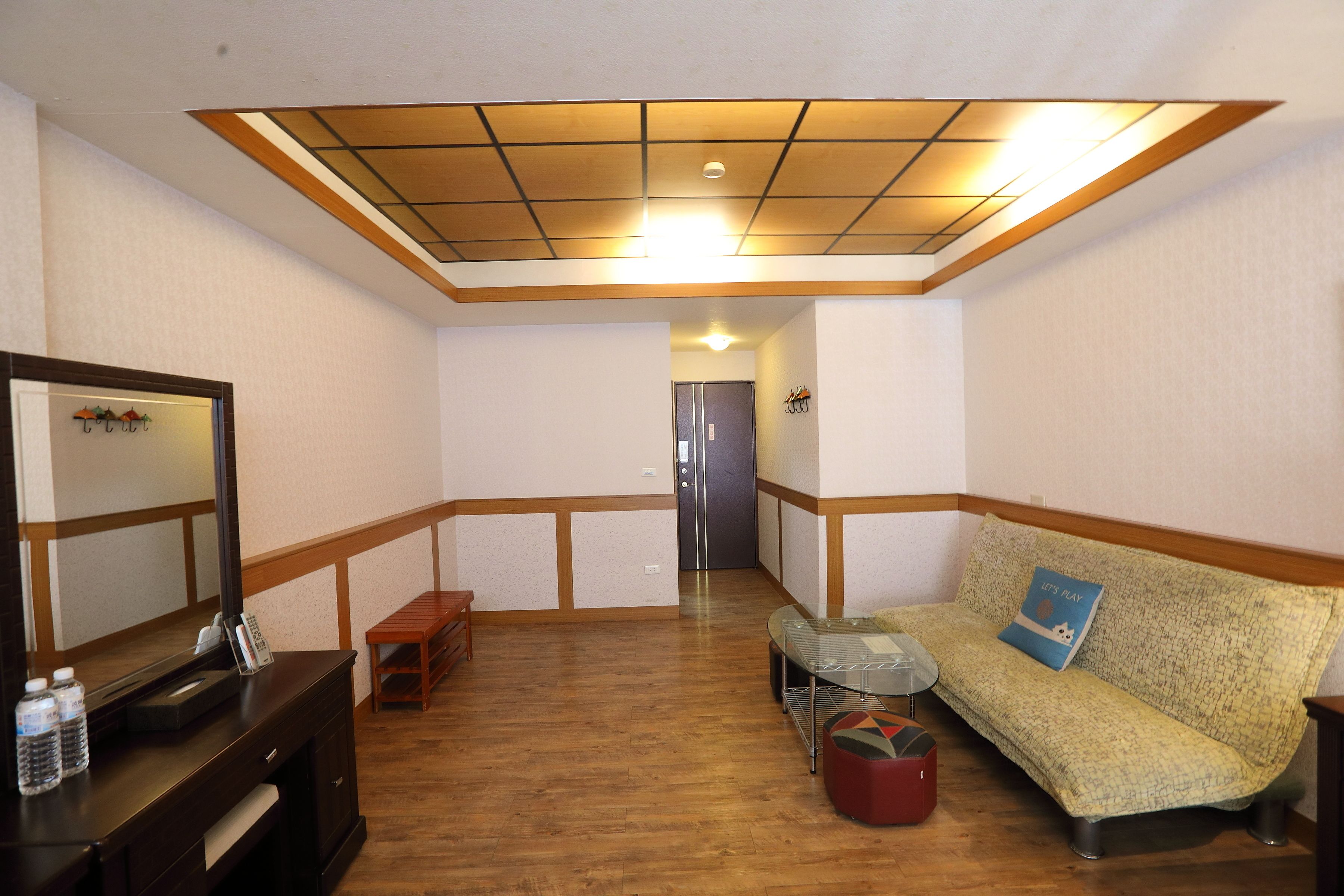 Une chambre d’hôtel avec un plancher en bois, un canapé avec un coussin, une petite table, un miroir et un bureau avec un téléphone et des bouteilles d’eau.