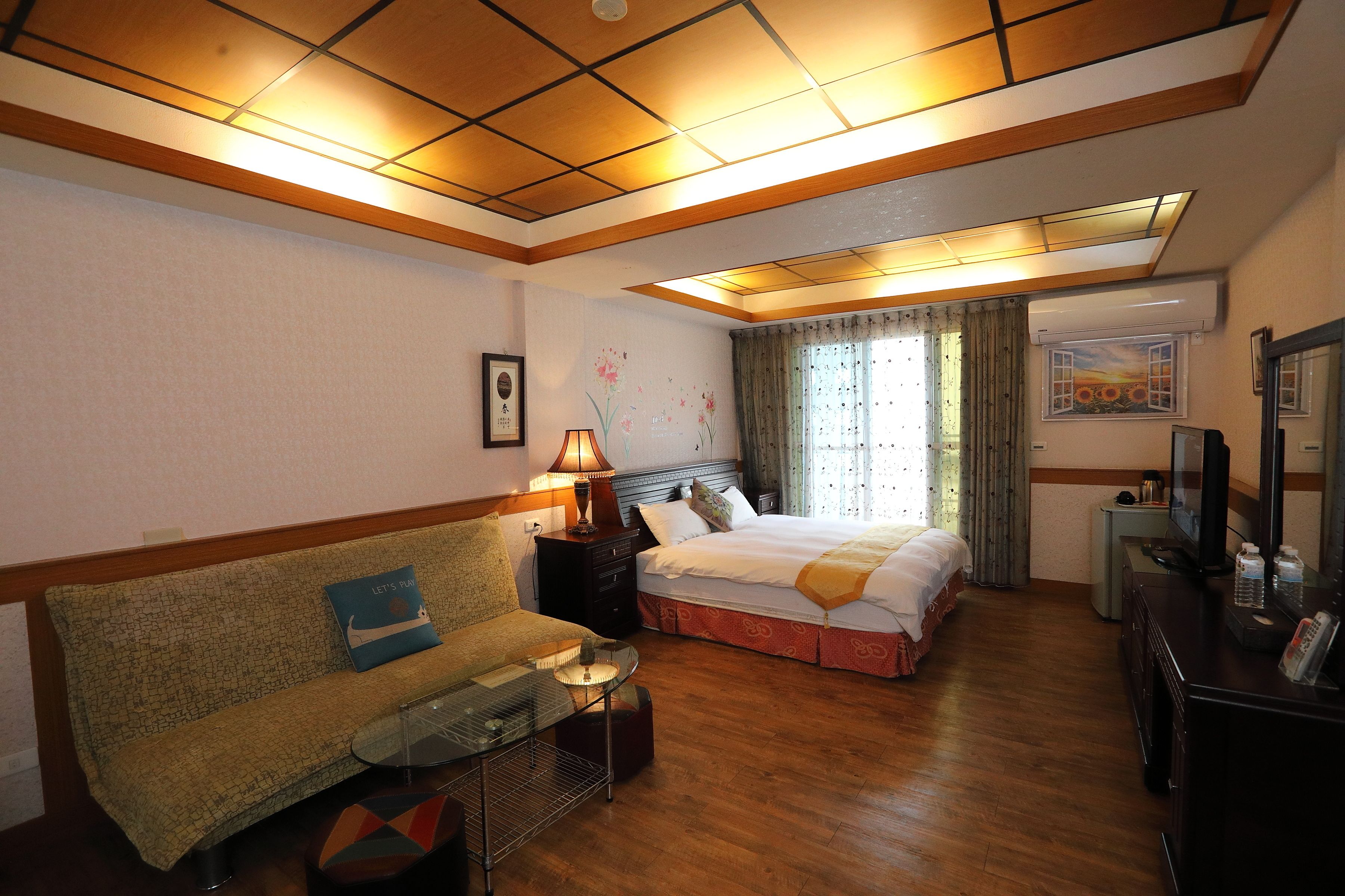 Quarto de hotel com sofá, cama, mesa de cabeceira, luminária, espelho e televisão.