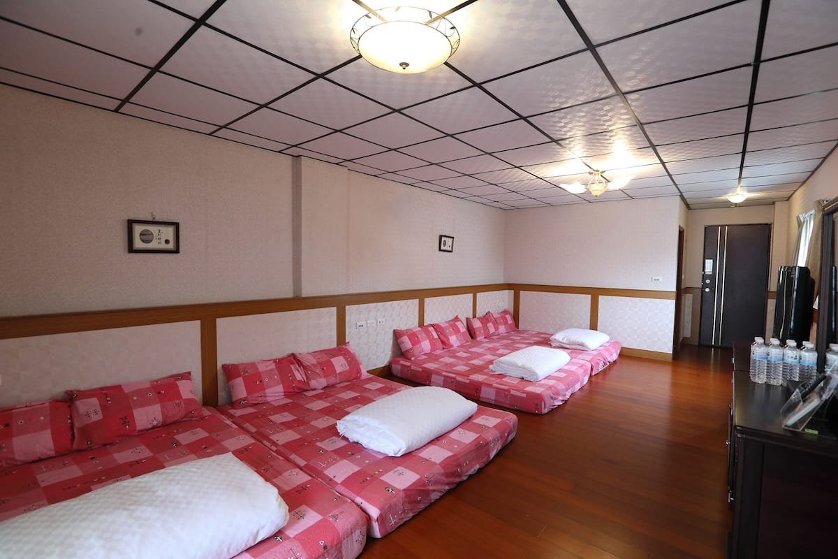 A hotel room with four beds arranged in a row, a wooden floor, and a ceiling with a grid pattern.