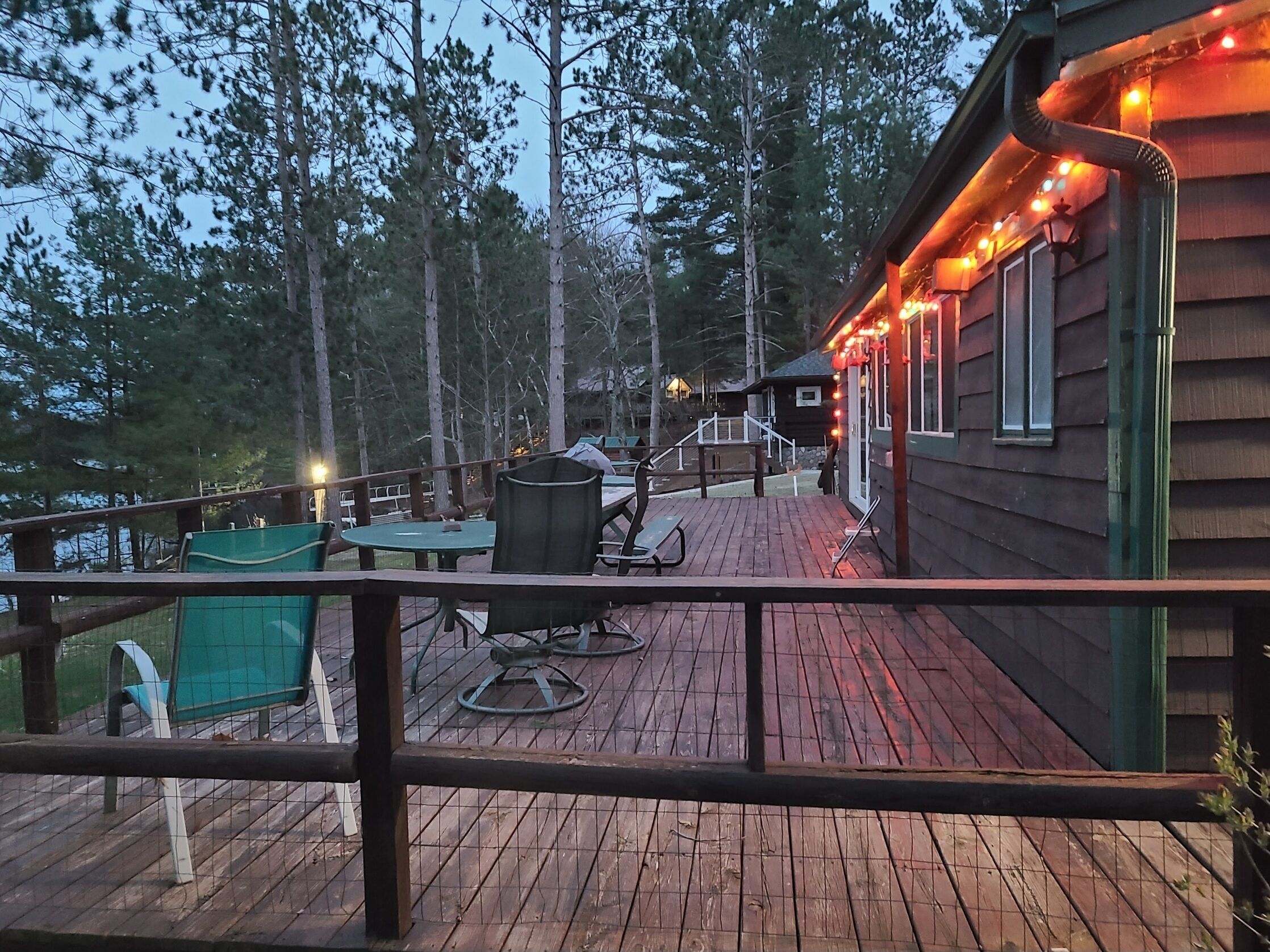 A wooden deck with outdoor seating, surrounded by trees, leading to a cabin with string lights.