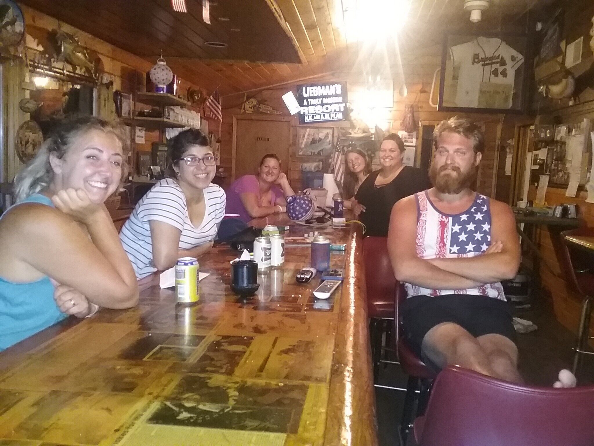 A group of people seated around a wooden bar with various items on it, including cans and a sign that reads "LIEBHMAN'S A TRULY MARIN RESORT."