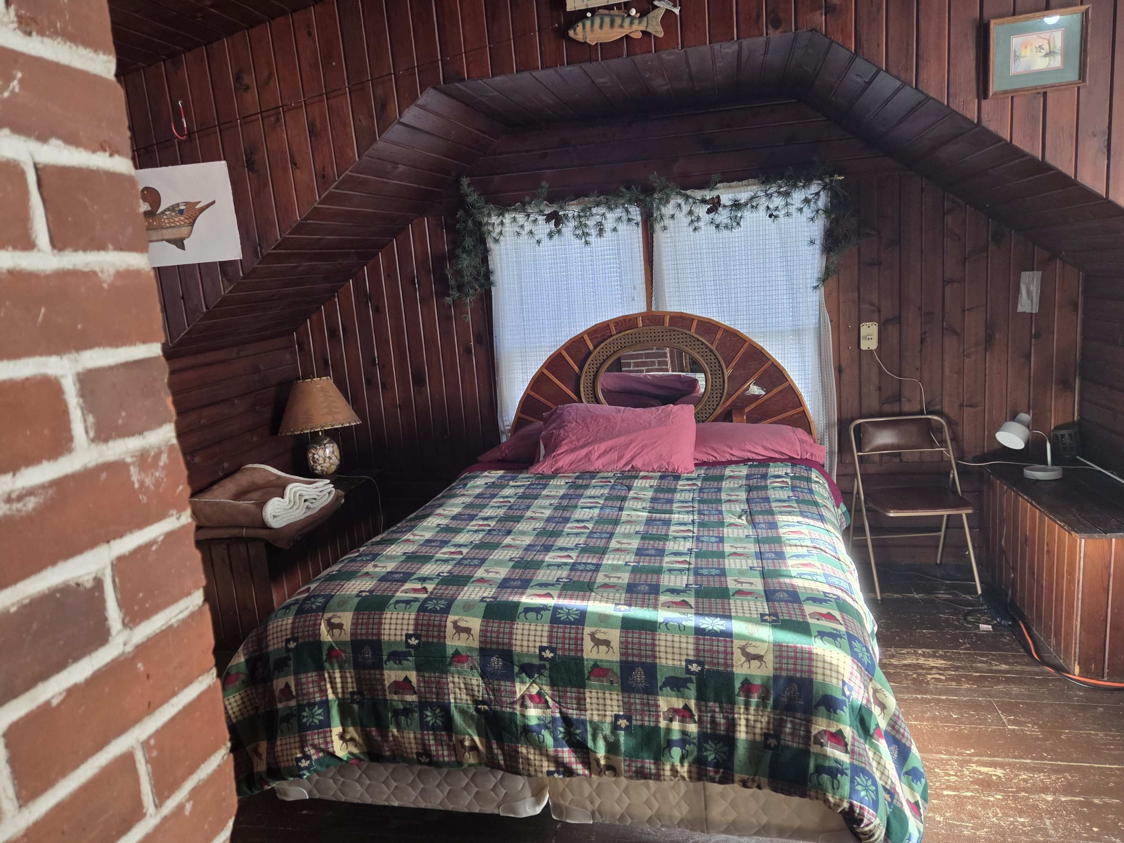 Un dormitorio acogedor en el ático con colcha de cuadros, cabecero de madera, mesita de noche con lámpara y pequeño escritorio con lámpara.