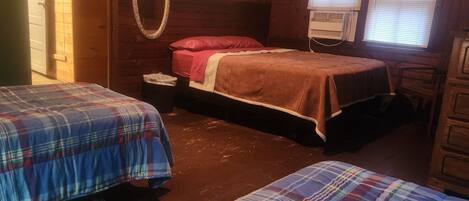 A cabin-style room with two beds, a wooden floor, a mirror, a heater, and a window with blinds.
