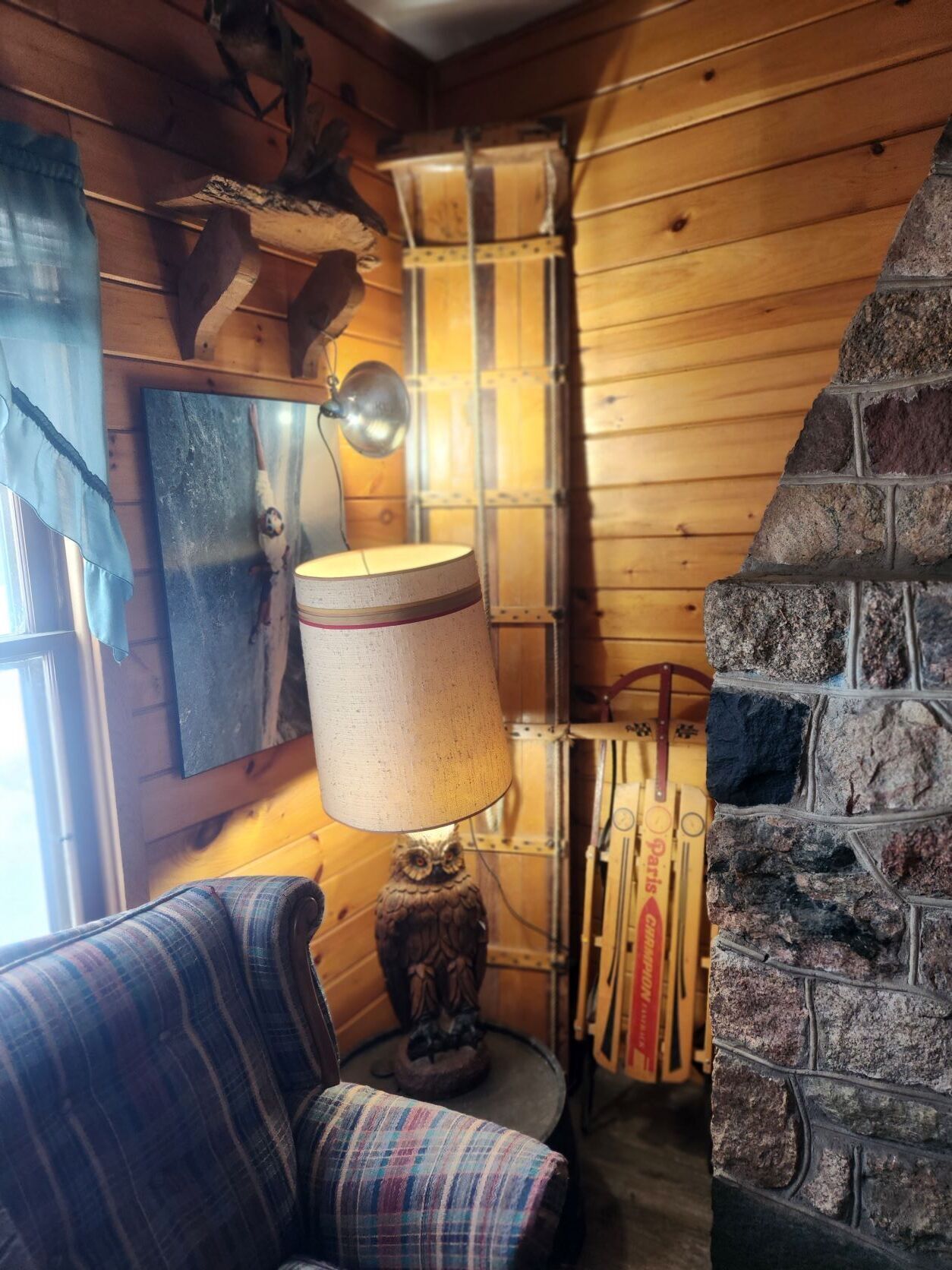 Un interior de cabaña acogedor con un sillón de cuadros, una lámpara, una pared de madera y una chimenea de piedra.