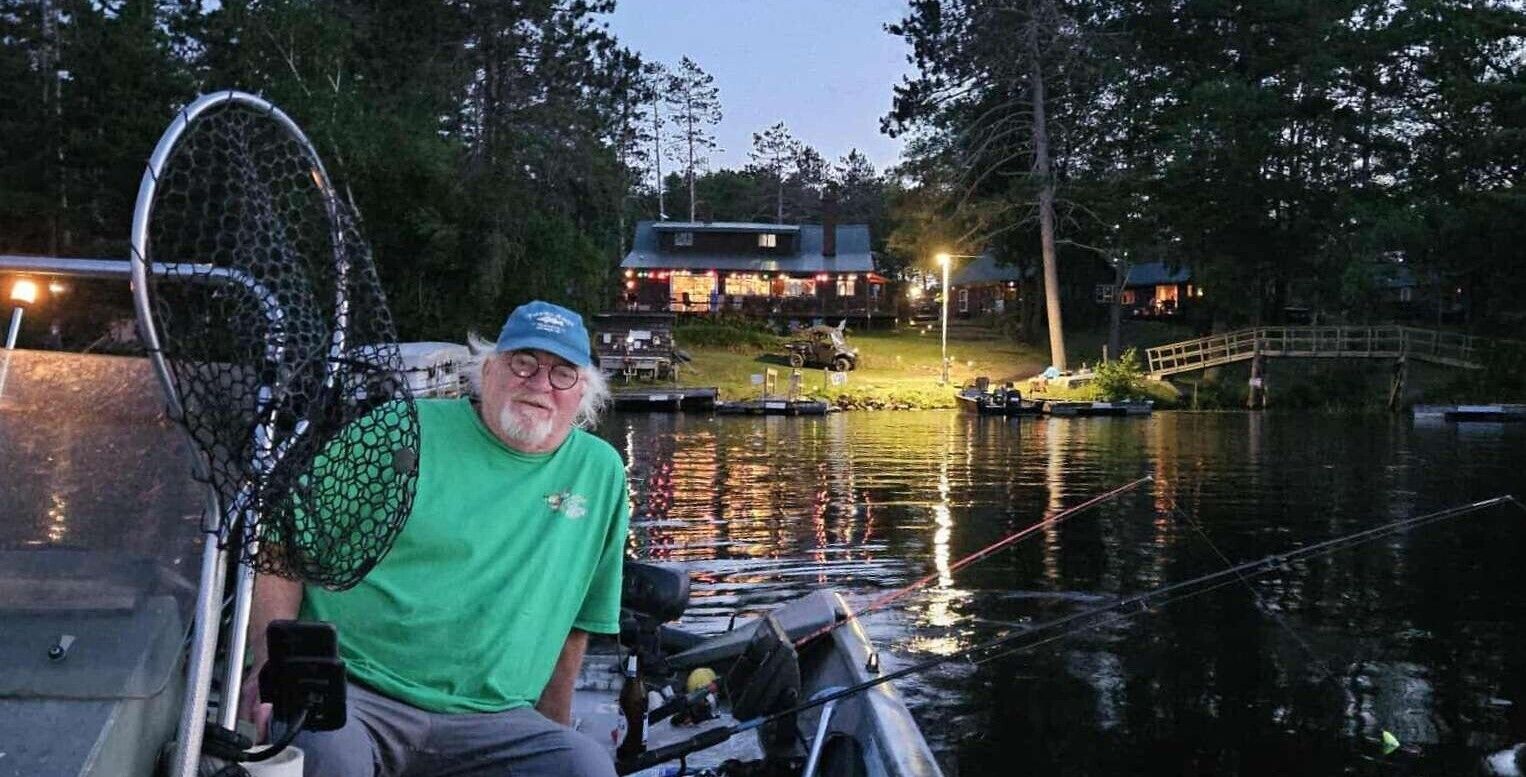 A person sitting on a dock by a calm body of water with boats, a house, and a bridge in the background.