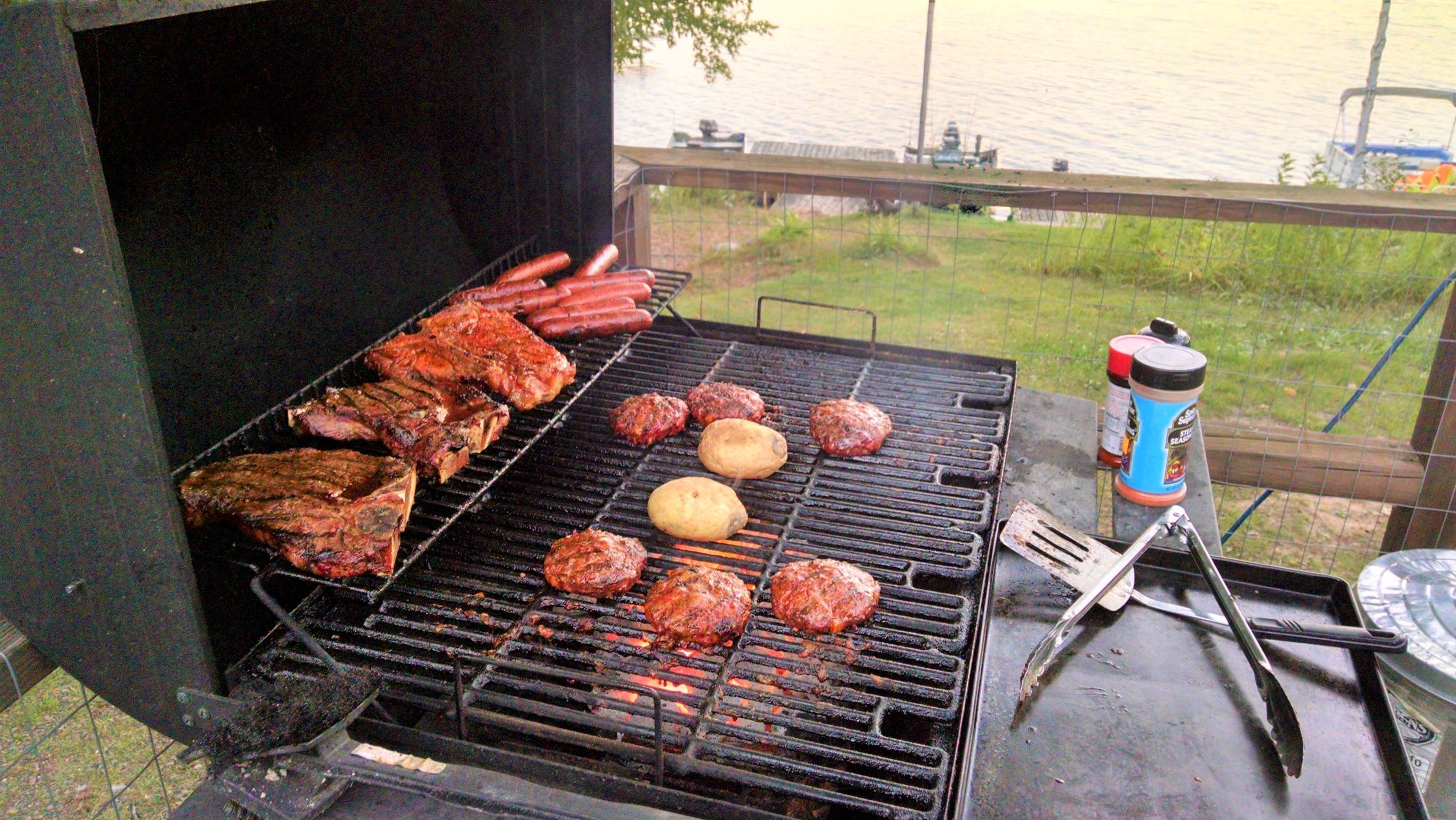 A grill with various meats and a potato, with a body of water and a fence in the background.