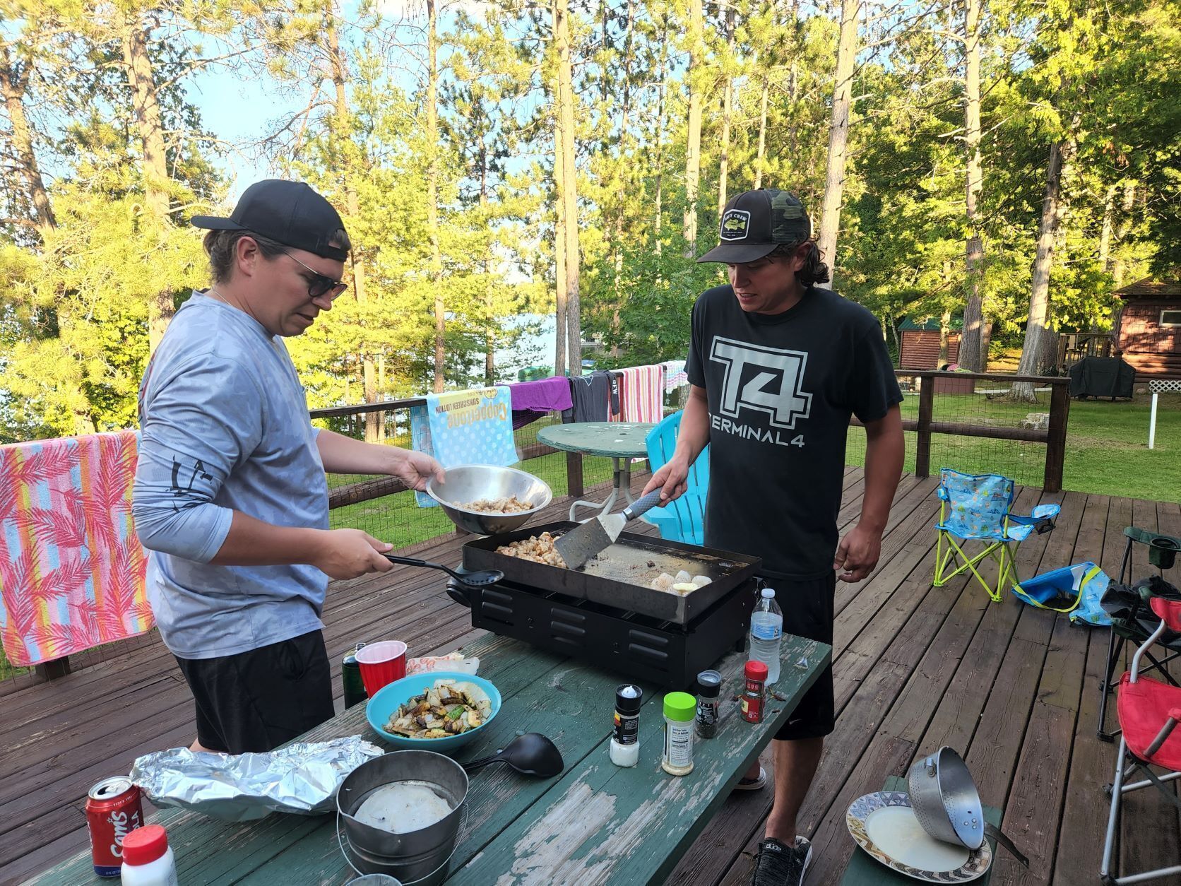 Two people cooking outdoors on a wooden deck with a grill, surrounded by trees.