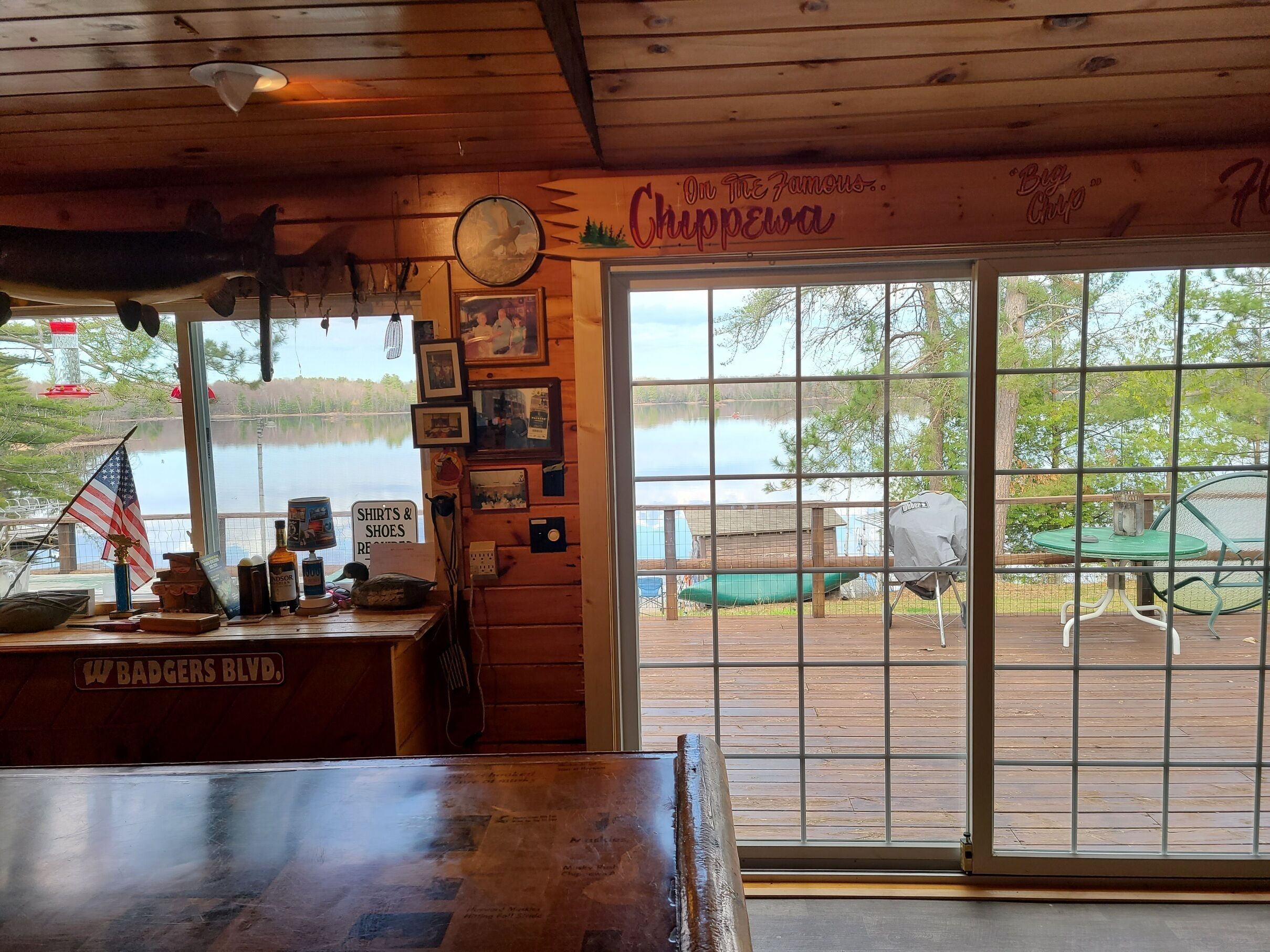 Un interior rústico con techo de madera, un reloj, cuadros enmarcados y vistas a un lago a través de amplias ventanas.