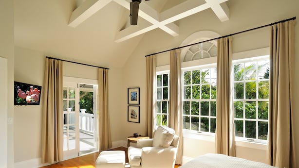 A bedroom with a large bed, a chair, a small table, and a TV. There are curtains, a ceiling fan, and a window with a view of greenery.