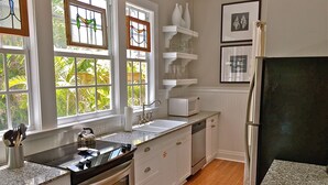 A kitchen with a stove, sink, and microwave. There are windows with curtains, a refrigerator, and shelves with glassware.