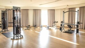A spacious gym with various exercise equipment, large windows with curtains, and a wooden floor. - The Energy Condo By AP (Cha-am)
