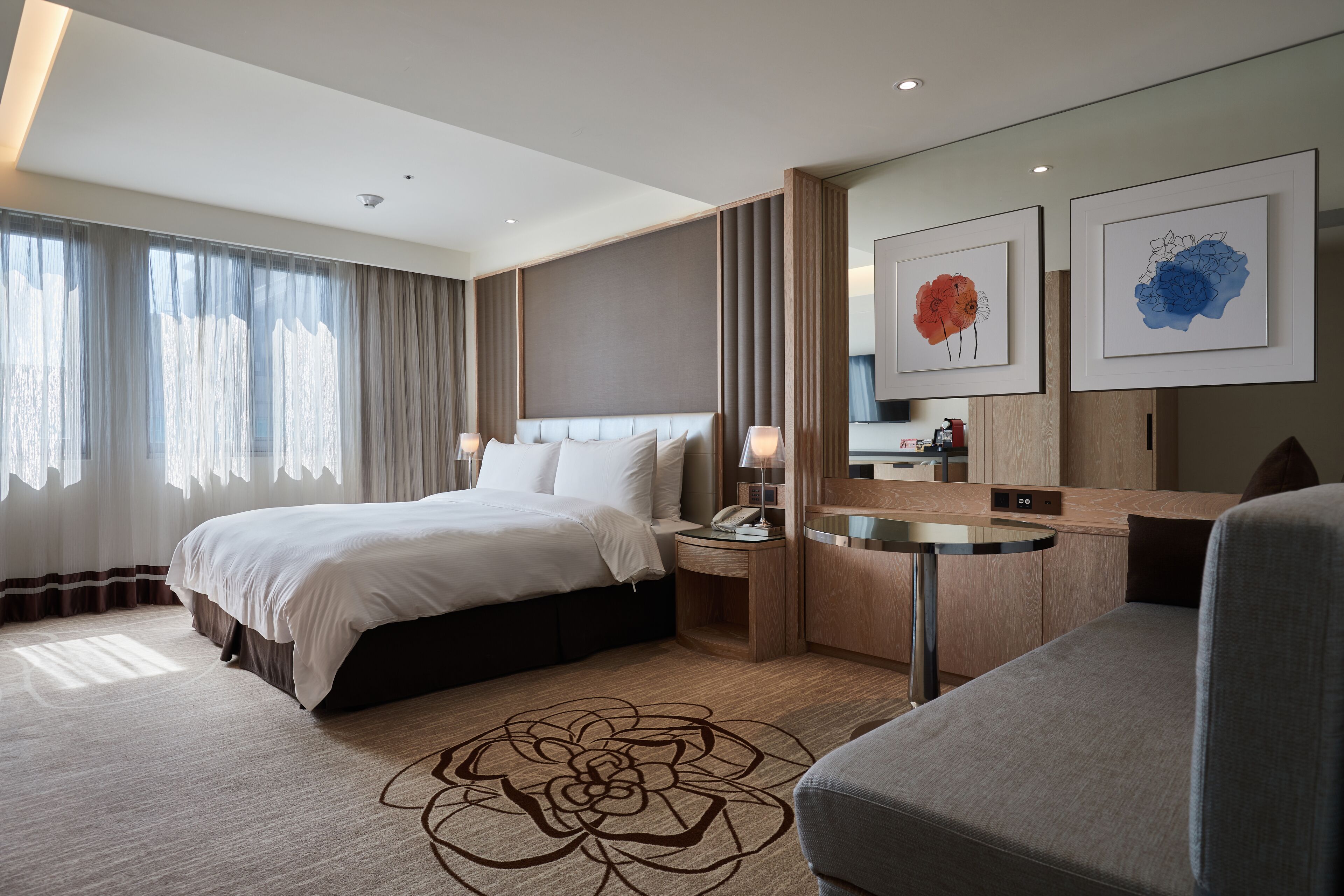 A modern hotel room with a large bed, a desk, and a chair. There is a window with curtains, a wall-mounted TV, and artwork above the desk.