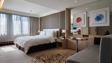 A modern hotel room with a large bed, a desk, and a chair. There is a window with curtains, a wall-mounted TV, and artwork above the desk.